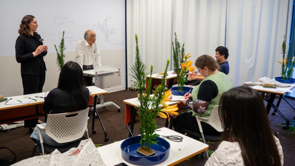 The flower arrangement instructor stands in front of the classroom and talks to the students, while iCLA students sit at their desks with their completed flower arrangements.