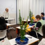 The flower arrangement instructor stands in front of the classroom and talks to the students, while iCLA students sit at their desks with their completed flower arrangements.