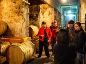 iCLA students are in a wine cellar surrounded by wine casks. They listen to the tour guide explain about Yamanashi wine making.