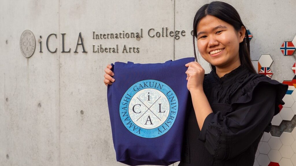 An iCLA student smiles and holds up a totebag by the iCLA sign in front of the college. On the totebag is her design of the iCLA logo which won the Memorabilia Logo Contest.