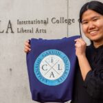 An iCLA student smiles and holds up a totebag by the iCLA sign in front of the college. On the totebag is her design of the iCLA logo which won the Memorabilia Logo Contest.